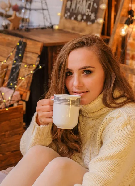 Portrait of young woman with cup of cocoa on bed at home. Minimalist christmas cozy decor on the background. Merry Christmas and Happy Holidays. Blonde, cute, woman in white sweater and warm scandinavian socks. New Year\'s Eve mood.