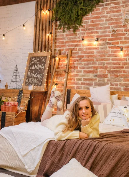 Young, smiling woman on bed at home. Minimalist christmas cozy decor on the background. Merry Christmas and Happy Holidays. Blonde, cute, woman in white sweater and warm scandinavian socks. New Year\'s Eve mood.