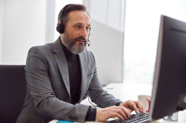 Kulaklıklı mutlu, olgun bir adam açık beyaz bir ofiste oturuyor.