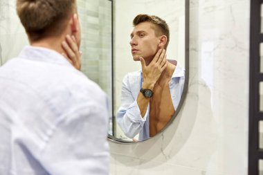 Modern bir banyoda, aynanın önünde duran beyaz, güzel bir adamın fotoğrafı.