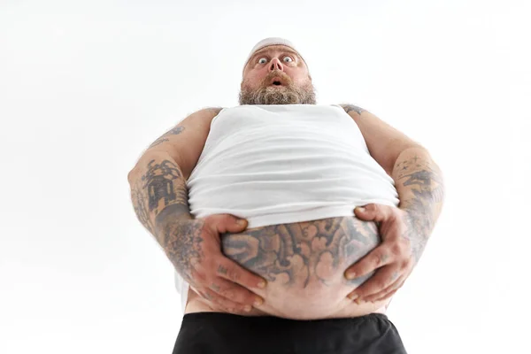 Dikke man met grote buik en tattoes in sportkleding houdt zijn maag met geschokte emotie — Stockfoto