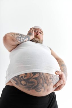 Koca göbekli ve spor giysilerindeki dövmeli şişman adam karnını şok edici bir duyguyla tutuyor.