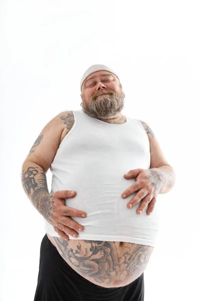 Boldog kövér férfi nagy pocakkal és tetovált lábujjakkal sportruhában tartja a gyomrát. — Stock Fotó