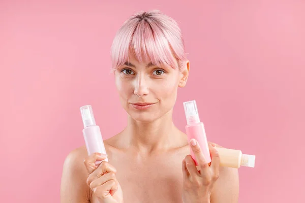 Attraktive junge Frau mit rosa Haaren blickt in die Kamera, hält drei Plastikflaschen mit Schönheitsprodukten in der Hand, posiert isoliert vor rosa Hintergrund — Stockfoto