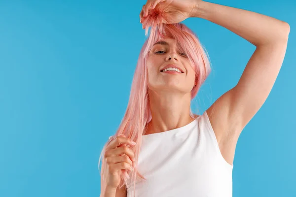 Glimlachend vrouwelijk model in wit shirt spelen met roze haar, wikkelen het rond haar gezicht terwijl poseren geïsoleerd over blauwe studio achtergrond — Stockfoto