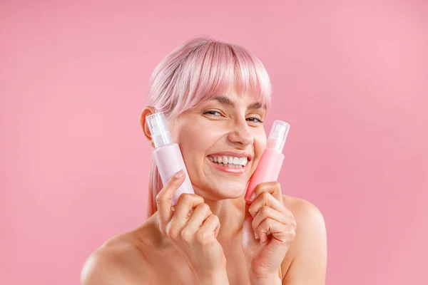 Zufriedene junge Frau mit rosa Haaren, die zwei Plastikflaschen mit Schönheitsprodukten vor ihrem Gesicht hält und isoliert auf rosa Hintergrund posiert — Stockfoto