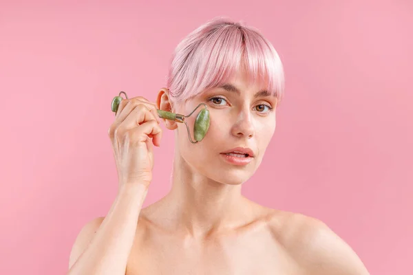 Retrato de mujer desnuda joven con pelo rosa usando rodillo facial de jade natural para el cuidado de la piel, posando aislado sobre fondo rosa — Foto de Stock