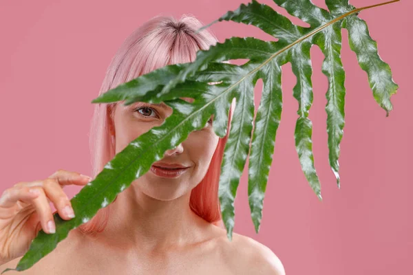 ピンクの髪をした若い女性がカメラを見て、緑の植物の葉の後ろに隠れて、ピンクのスタジオの背景に隔離されたポーズ — ストック写真