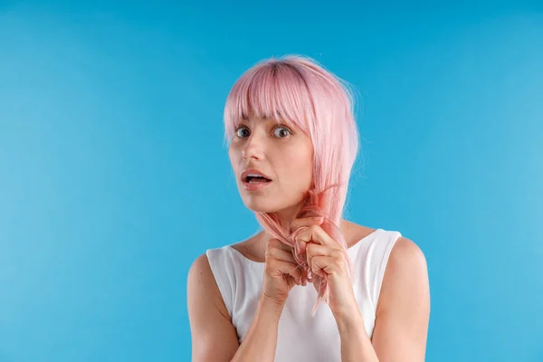 Portret zaskoczonej modelki bawiącej się różowymi włosami, dotykającej jej podczas pozowania na niebieskim tle studia — Zdjęcie stockowe