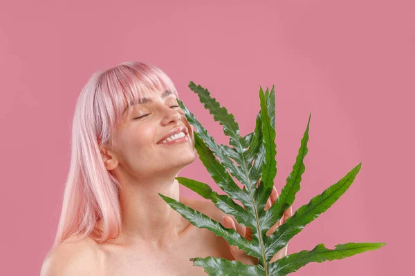 Retrato de mujer complacida con el pelo rosado sonriendo con los ojos cerrados, sosteniendo la hoja de la planta cerca de su cara, posando aislada sobre fondo rosa estudio — Foto de Stock