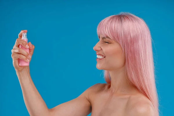 Portret van vrolijke vrouw met roze haar met behulp van spray in plastic fles over haar gezicht met gesloten ogen, poseren geïsoleerd over blauwe studio achtergrond — Stockfoto