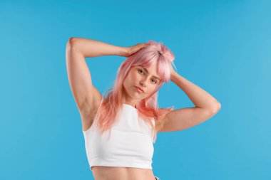 Beyaz gömlekli çekici bir kadın kameraya bakıyor. Pembe saçlarını düzeltirken mavi stüdyo arka planında yalnız poz veriyor.