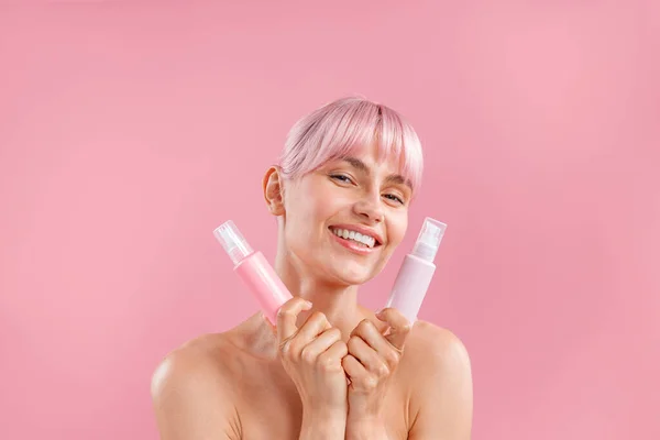 Porträt einer glücklichen jungen Frau mit rosa Haaren, die zwei Plastikflaschen mit Schönheitsprodukten hält und isoliert auf rosa Hintergrund posiert — Stockfoto
