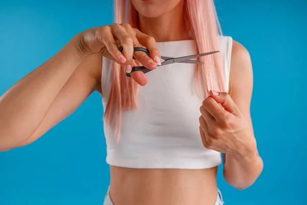 Schnappschuss einer Frau mit rosa Haaren, die sich mit einer Schere die Haare schneidet, isoliert vor blauem Studiohintergrund — Stockfoto