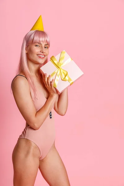 Glad ung kvinna med rosa hår bär baddräkt och fest hatt hålla söt presentförpackning, stående isolerad över rosa bakgrund — Stockfoto
