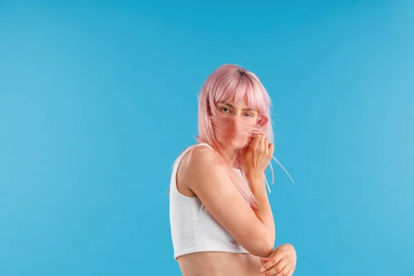 Aantrekkelijke vrouw in wit shirt kijken naar camera, verbergen haar gezicht met roze haar terwijl poseren geïsoleerd over blauwe studio achtergrond — Stockfoto