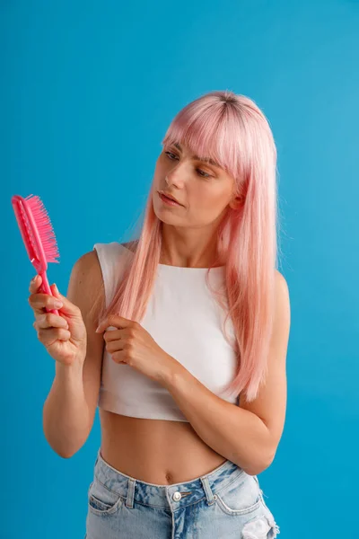 Neugierige Frau mit glatten natürlichen langen rosafarbenen Haaren schaut auf Haarkamm, während sie isoliert vor blauem Studiohintergrund steht — Stockfoto