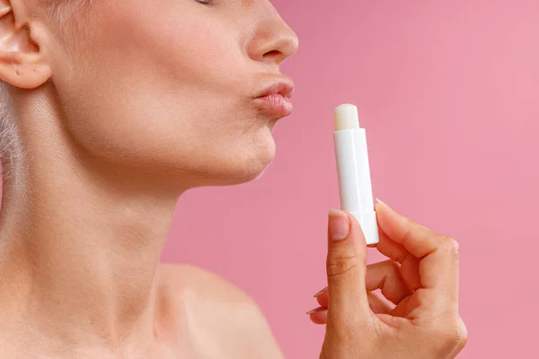 Close up shot of woman holding lip balm near her lips and making kissing gesture, posing isolated over pink background