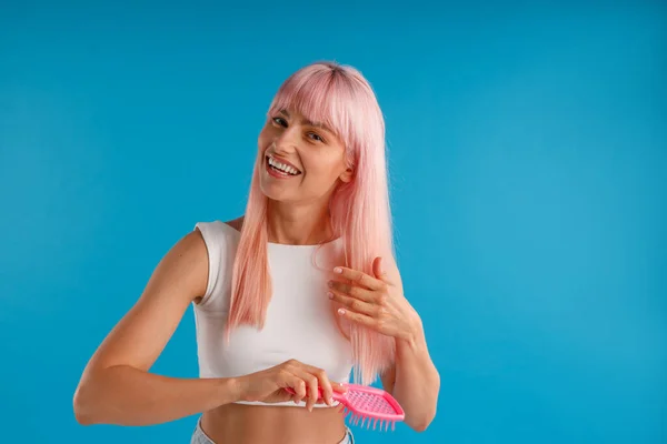 Glada kvinna borsta hennes släta naturliga lång rosa hår med hår kam samtidigt poserar isolerad över blå studio bakgrund — Stockfoto