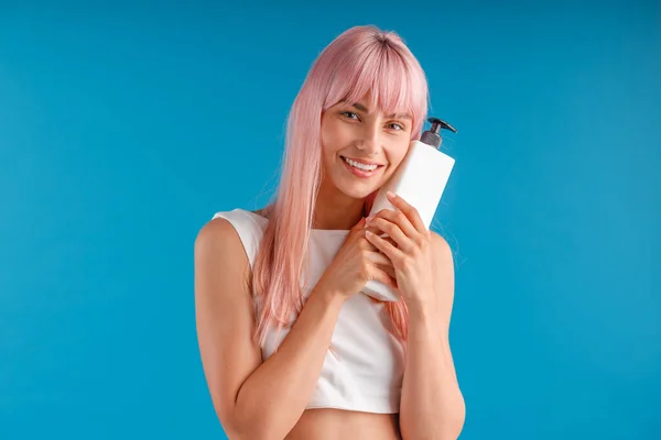 Щаслива жінка з гладким рожевим волоссям тримає і обіймає пляшку насоса з маскою для волосся або шампунем, позує ізольовано на синьому студійному фоні — стокове фото