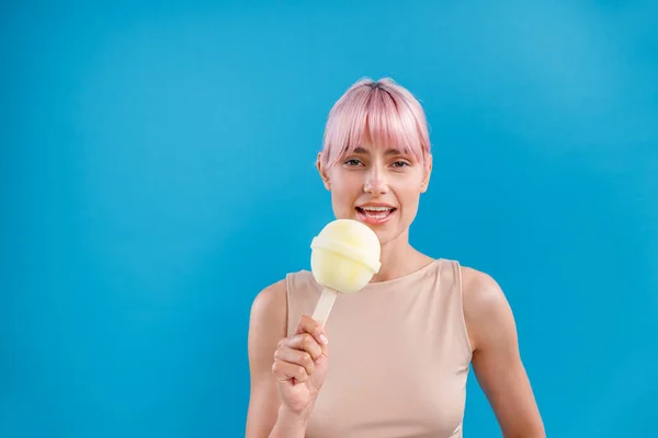 Porträtt av attraktiv kvinna med rosa hår tittar på kameran och håller en stor klubba godis glass framför henne, poserar isolerad över blå bakgrund — Stockfoto