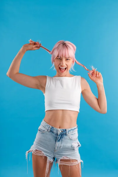 Aufgeregte junge Frau in Freizeitkleidung lächelt in die Kamera und spielt mit ihren rosa Haaren, während sie isoliert vor blauem Studiohintergrund steht — Stockfoto