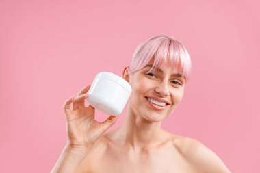 Pembe saçlı gülümseyen kadının portresi pembe arka planda izole edilmiş duş sonrası nemlendirici vücut losyonuyla beyaz kavanozunu tutuyor.