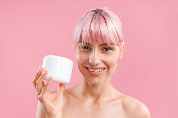 Portrait of smiling woman with pink hair holding white jar with nourishing body cream after shower isolated over pink background