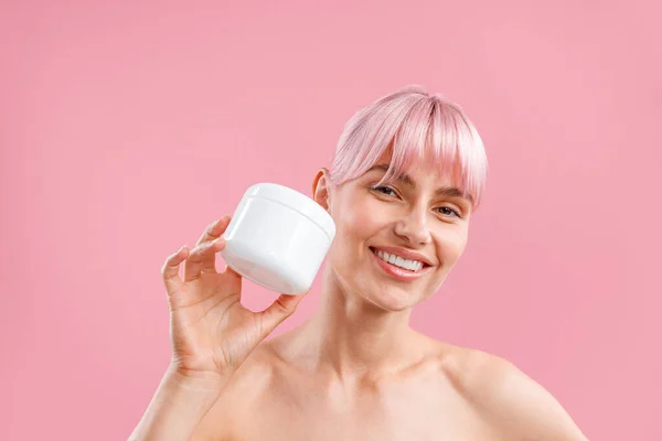 Portret van een lachende vrouw met roze haar houden witte pot met hydraterende bodylotion na douche geïsoleerd over roze achtergrond — Stockfoto
