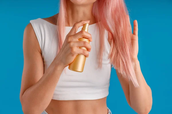 Nahaufnahme der Hände einer jungen Frau, die ihr glattes rosafarbenes Haar, isoliert vor blauem Studiohintergrund stehend, mit Feuchtigkeitsspray versorgt und anwendet — Stockfoto