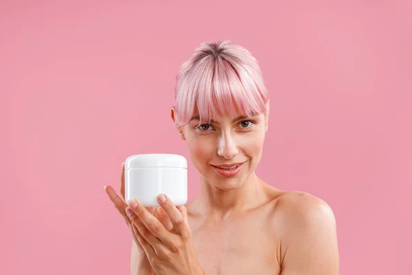 Porträt einer Frau mit rosafarbenen Haaren, die in die Kamera blickt und ein weißes Glas mit feuchtigkeitsspendender Körperlotion nach der Dusche isoliert vor rosa Hintergrund zeigt — Stockfoto