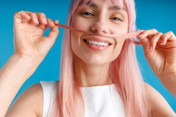 Close-up portret van gelukkige jonge vrouw met natuurlijke lange roze geverfd haar met een streng van haar als een snor en glimlachen op de camera, poseren geïsoleerd over blauwe studio achtergrond — Stockfoto