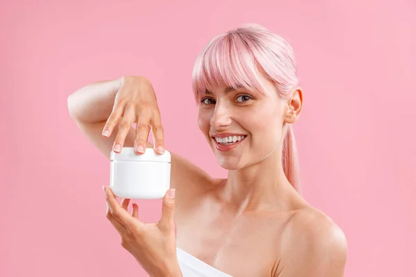 Portret van jonge vrouw glimlachen op camera, met witte pot met hydraterende bodylotion na douche geïsoleerd over roze achtergrond — Stockfoto