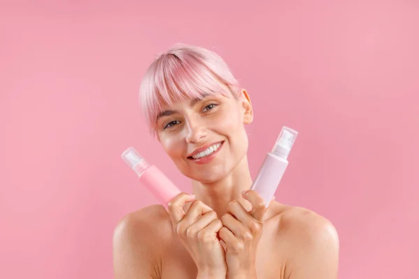 Porträt einer schönen jungen Frau mit rosa Haaren, die zwei Plastikflaschen mit Schönheitsprodukten hält und isoliert auf rosa Hintergrund posiert — Stockfoto