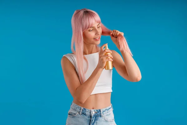Vårdande ung kvinna ler och tillämpa fuktgivande spray till hennes släta rosa hår stående isolerad över blå studio bakgrund — Stockfoto