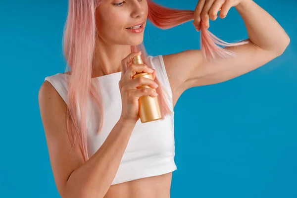 Nahaufnahme einer jungen Frau, die Feuchtigkeitsspray auf ihre rosa Haare aufträgt, während sie isoliert vor blauem Studiohintergrund steht — Stockfoto