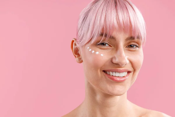 Retrato de mujer sonriente con cabello rosa y crema nutritiva aplicada en su cara como puntos aislados sobre fondo rosa — Foto de Stock