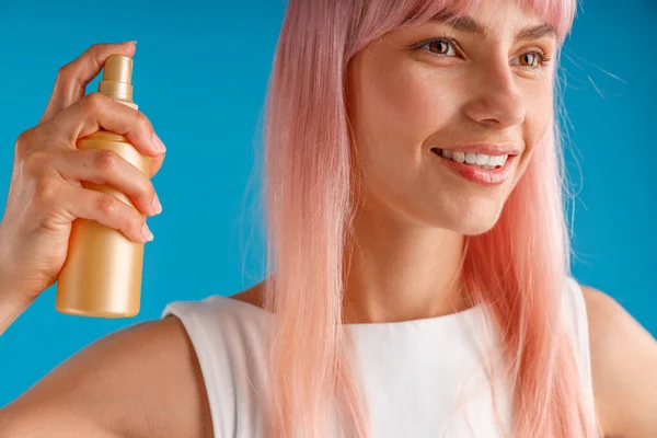 Close-up portret van mooie jonge vrouw met roze haar glimlachen opzij en houden fles hydraterende spray, poseren geïsoleerd over blauwe studio achtergrond — Stockfoto