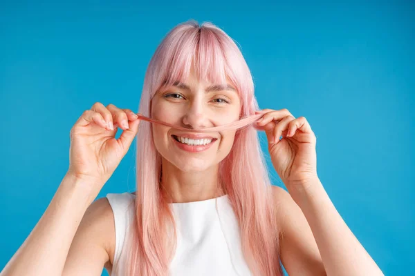 Portrait of happy young woman with natural long pink dyed hair holding a strand of hair as a moustache and smiling at camera, posing isolated over blue studio background — Stock Photo, Image