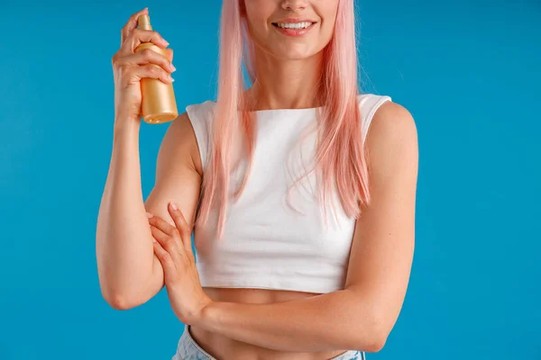 Schnappschuss einer jungen Frau mit rosa Haaren, die lächelt und eine Flasche feuchtigkeitsspendendes Spray in der Hand hält, während sie isoliert vor blauem Studiohintergrund steht — Stockfoto