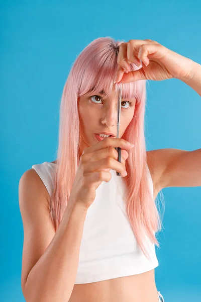 Schattige jonge vrouw met roze haar knippen franje zelf met behulp van schaar, staande geïsoleerd over blauwe studio achtergrond — Stockfoto