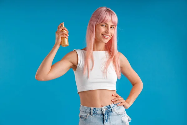 Gelukkige jonge vrouw met roze haar glimlachen en houden fles van haar conditioner, hydraterende spray terwijl staande geïsoleerd over blauwe studio achtergrond — Stockfoto