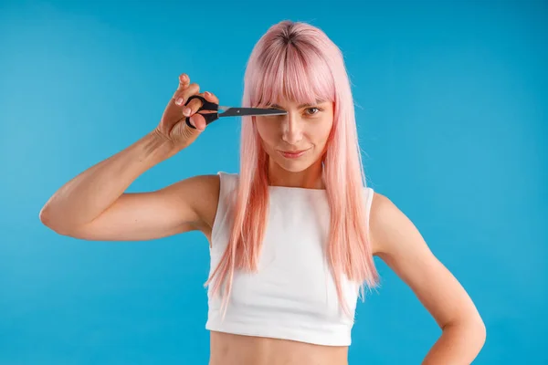 Ernste junge Frau mit rosafarbenen Haaren, die eine Schere vor dem Gesicht hält und isoliert vor blauem Studiohintergrund posiert — Stockfoto