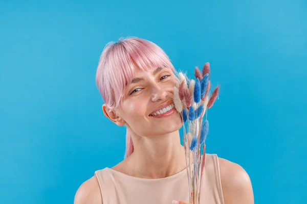 Porträtt av vacker kvinna med rosa hår ler mot kameran, håller torkade pampas gräs i handen, poserar över blå studio bakgrund — Stockfoto