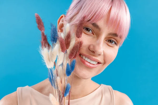 Primer plano de la hermosa mujer con el pelo rosa sonriendo a la cámara, sosteniendo flores secas en su mano, posando sobre el fondo azul del estudio. Tallo de caña, composición de flores secas de estilo nórdico artificial — Foto de Stock