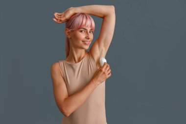 Gri stüdyo arka planında izole edilmiş, terleme önleyici kullanırken kameraya gülümseyen bej elbiseli, pembe saçlı mutlu genç kadın.