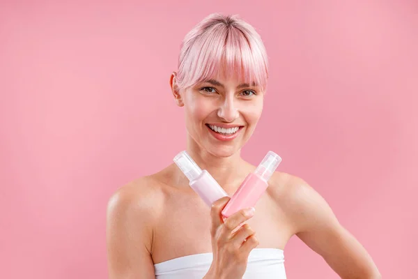 Opgewonden jonge vrouw met roze haar glimlachend en met twee plastic spay flessen met schoonheidsproducten, poseren geïsoleerd over roze achtergrond — Stockfoto