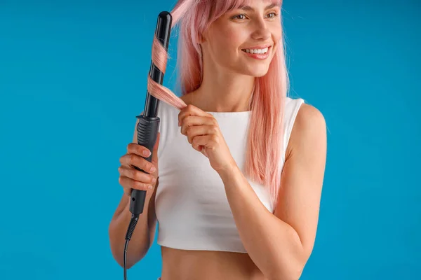 Schöne lächelnde Frau Styling rosa lange glatte Haare mit Lockenstab, steht isoliert über blauem Studiohintergrund — Stockfoto