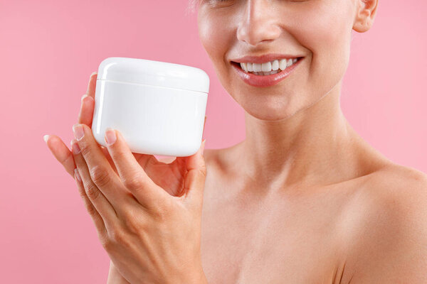 Close up shot of young woman holding white jar with moisturizing body lotion after shower isolated over pink background