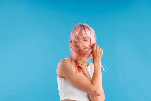 Glimlachende vrouw model in wit shirt spelen met gladde rechte roze haar, verbergen haar gezicht terwijl poseren geïsoleerd over blauwe studio achtergrond — Stockfoto